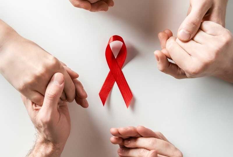 Manos formando un círculo alrededor de un listón rojo, símbolo de conciencia y apoyo en la lucha contra el VIH.